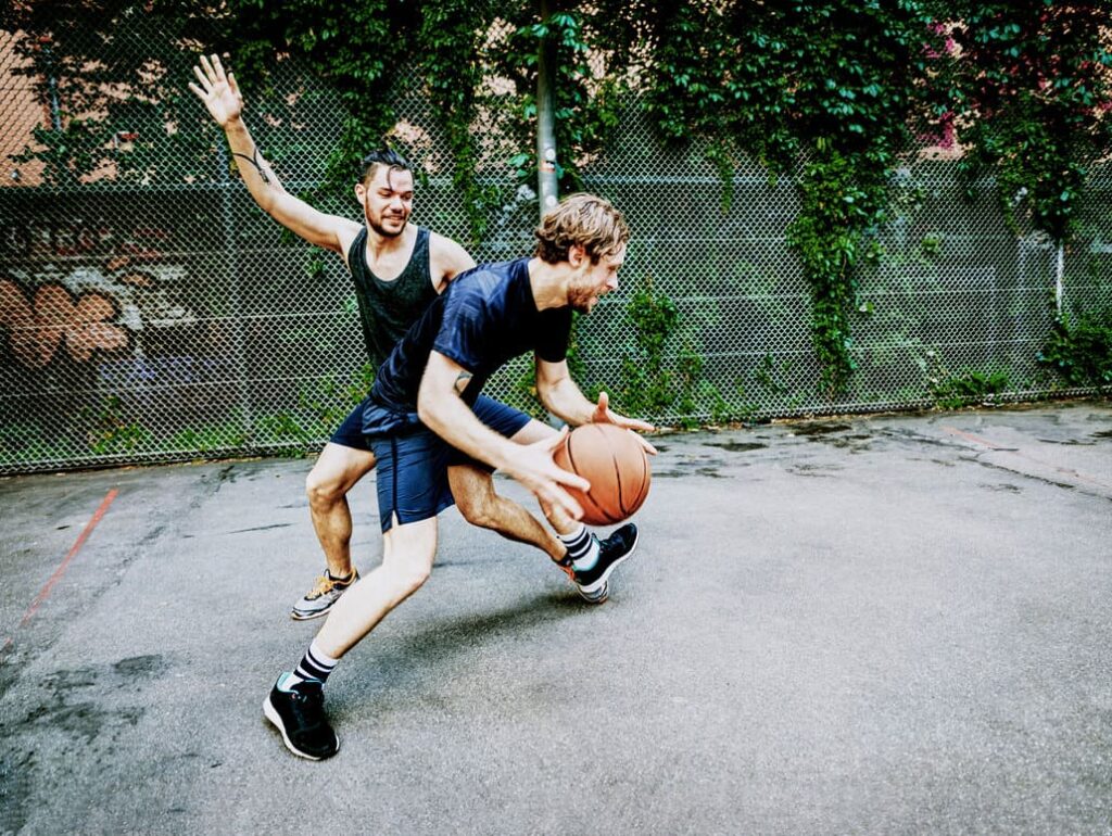 Auf dem Bild sind zwei Männer zu sehen, die vor einer grünen Wand Basketball spielen.