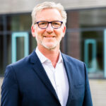 Porträtfoto von Matthias Albrecht, einem Mann mit grauem Haar und Brille, der in die Kamera lächelt. Er trägt ein helles Hemd und ein Sakko. Bildnachweis: Lena Sielaff. Matthias Albrecht ist Facharzt für Kinder- und Jugendmedizin und Geschäftsführer bei KLUG, wo er den Aufbau eines Kompetenzzentrums für klimaresiliente Gesundheitseinrichtungen verantwortet. Zuvor leitete er 15 Jahre das Evangelische Krankenhaus Hubertus in Berlin – das erste vom BUND zertifizierte Krankenhaus Deutschlands. Er ist Mitglied der Arbeitsgruppe „Klimawandel
